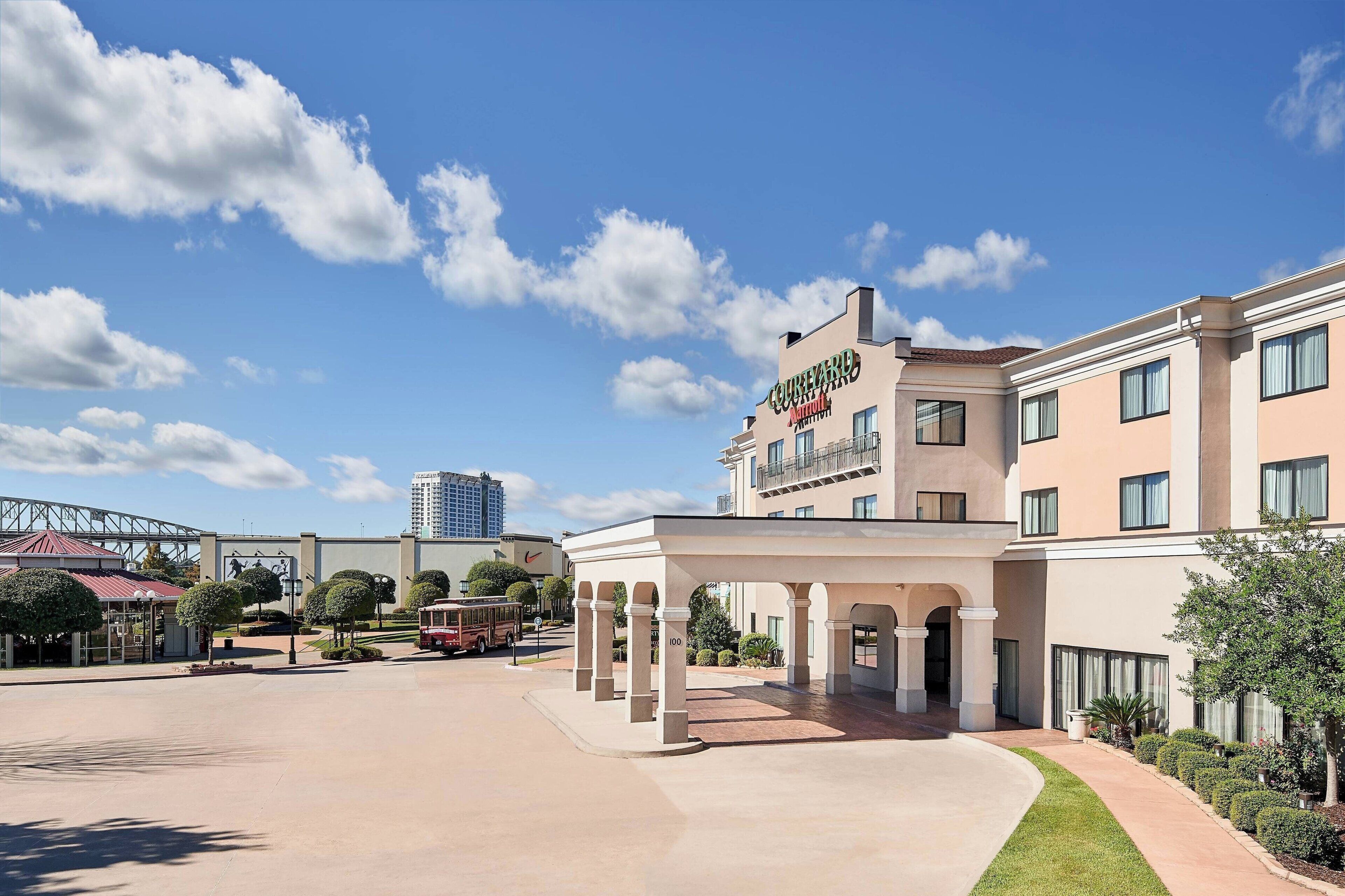 A hotel with a covered entrance, a parking area, and a view of a bridge and tall buildings in the background.