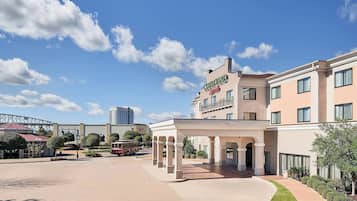 A hotel with a covered entrance, a parking area, and a view of a bridge and tall buildings in the background.