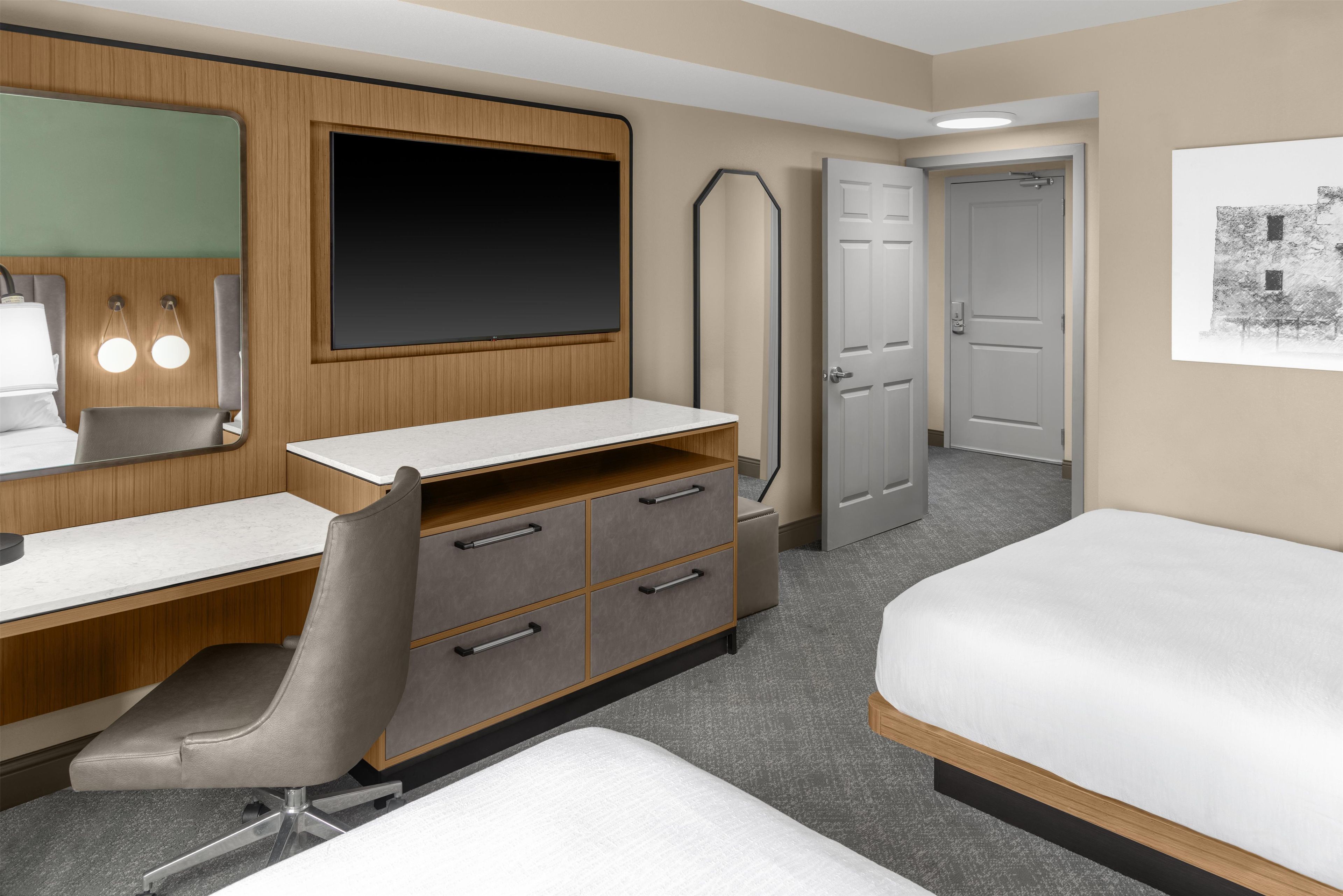 Une chambre d’hôtel comprenant un lit, un bureau avec des tiroirs, une chaise, un grand miroir, un téléviseur à écran plat et une porte.