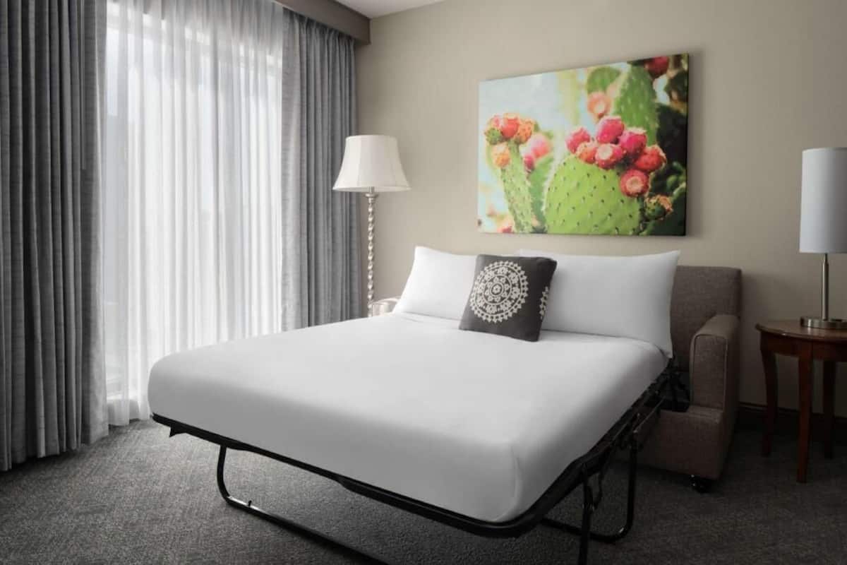 A hotel room with a bed, a lamp, a chair, a painting, and a window with curtains.