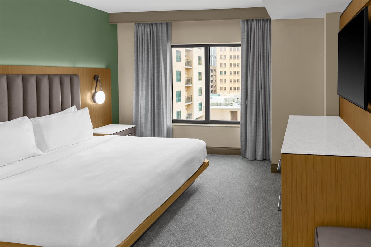A hotel room with a large bed, a bedside table, a TV, a window with curtains, and a view of buildings.