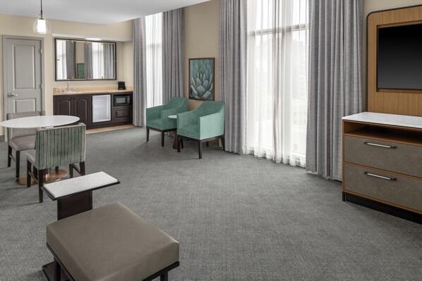 A hotel room with a kitchenette, a dining area with a table and chairs, a seating area with two chairs, a large window with curtains, and a television. - Embassy Suites San Antonio Riverwalk-Downtown (San Antonio)