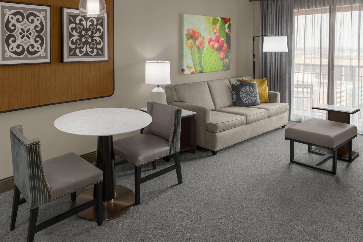 A hotel room with a round dining table, two chairs, a sofa, a bench, a lamp, and a cactus artwork on the wall.