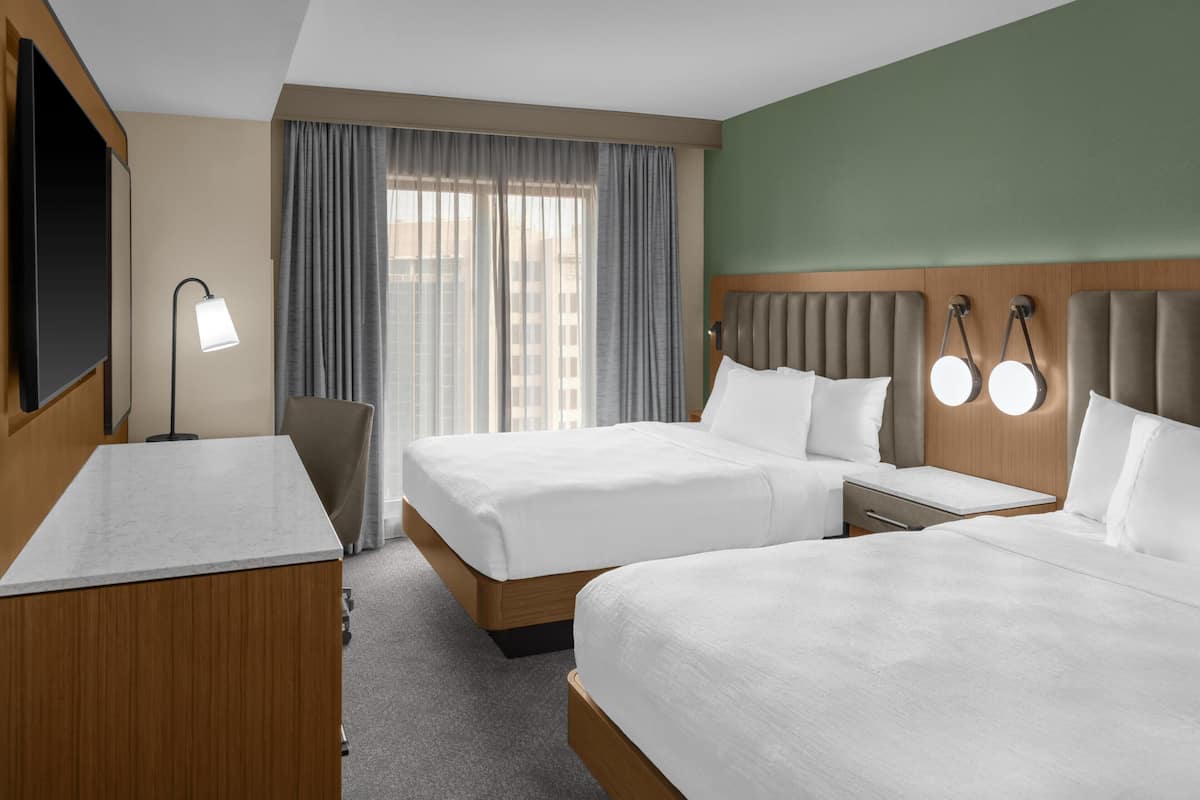 A hotel room with two beds, a desk, a chair, a TV, and a window with curtains.