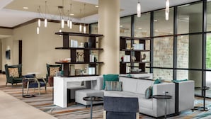 A modern lobby with a sectional sofa, armchairs, a coffee table, a bookshelf, and a large window with a stone wall. - Embassy Suites San Antonio Riverwalk-Downtown (San Antonio)