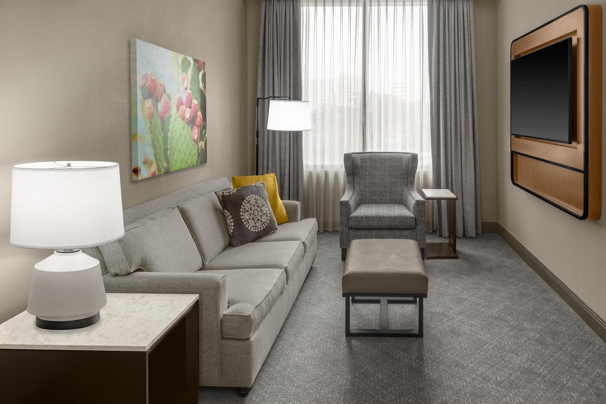 A hotel room with a sofa, armchair, ottoman, TV, lamp, and artwork on the wall.