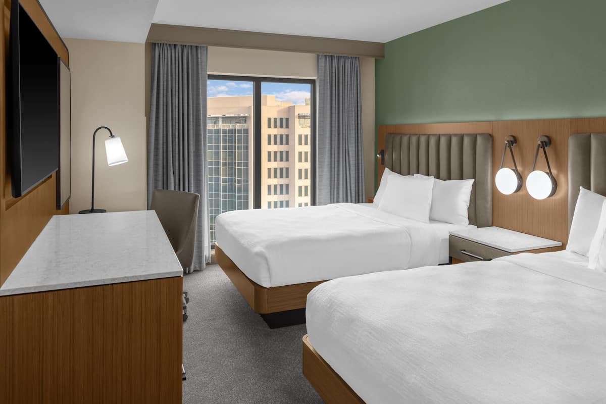 A hotel room with two beds, a desk, a chair, a TV, a lamp, and a view of buildings through the window.