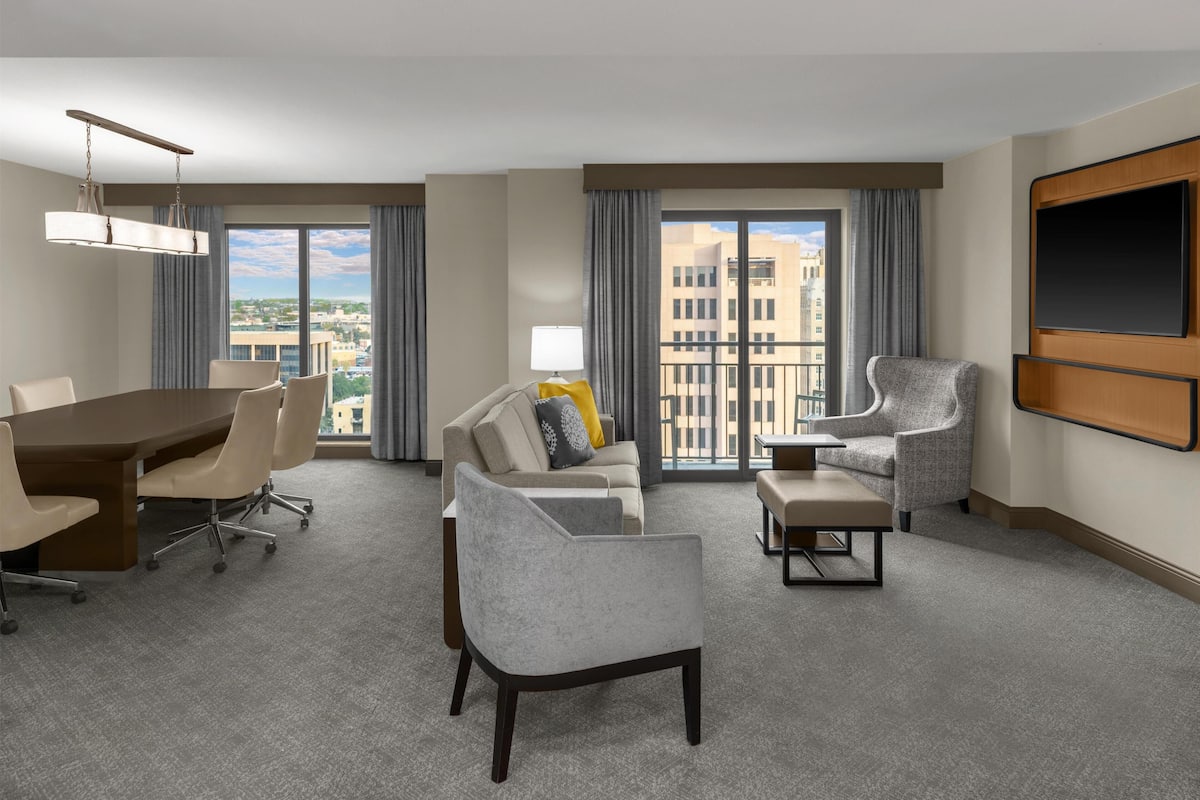 A hotel room with a dining table, two sofas, a TV, and large windows with a city view.