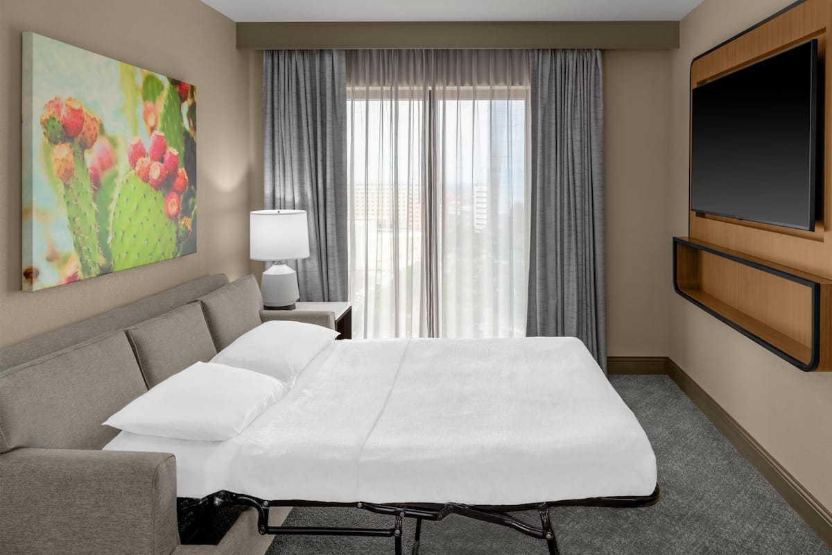 A hotel room with a bed, a sofa, a TV, a window with curtains, and a painting on the wall.