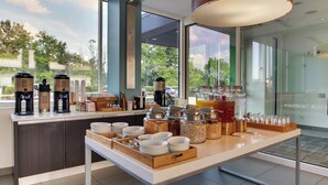 A breakfast bar with various food items and beverages, a coffee machine, and a large window offering a view of the outdoors.