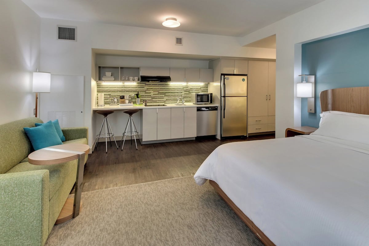Quarto de hotel com uma cama, um sofá verde, uma pequena mesa redonda e uma cozinha compacta com micro-ondas, frigorífico e banquetas.