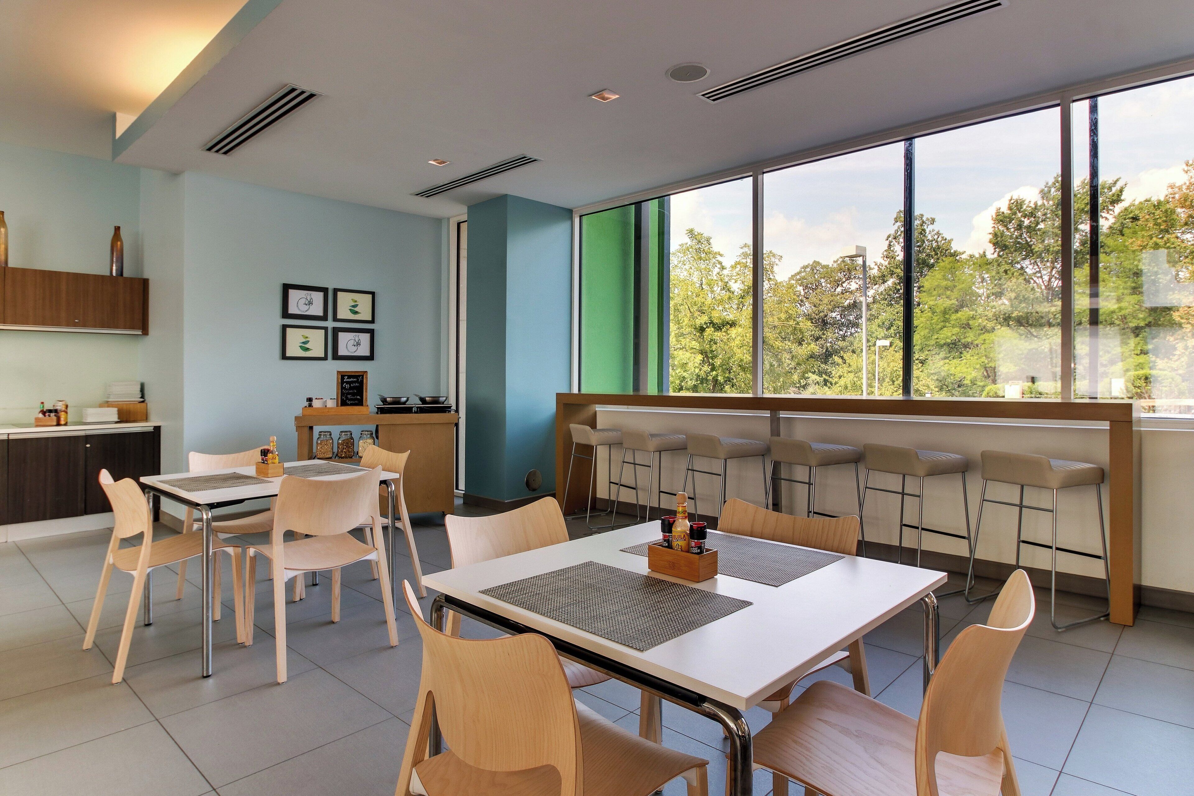 A modern dining area with a large window, a kitchen in the background, and a seating arrangement for guests.