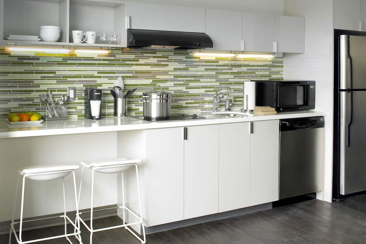 Cozinha moderna com armários brancos, geladeira de aço inoxidável, micro-ondas e painel de azulejos.