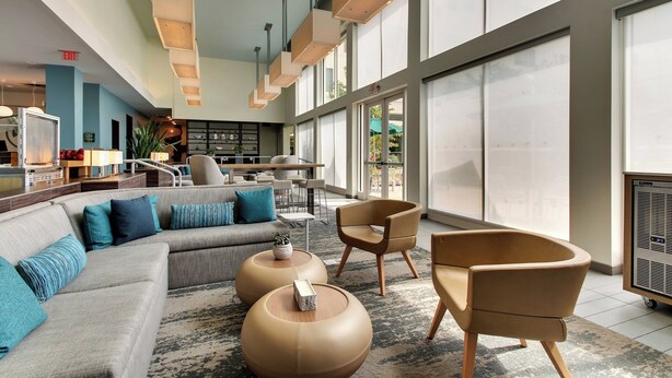 A spacious lobby with a large sofa, armchairs, and a coffee table. There are large windows and a reception desk in the background.