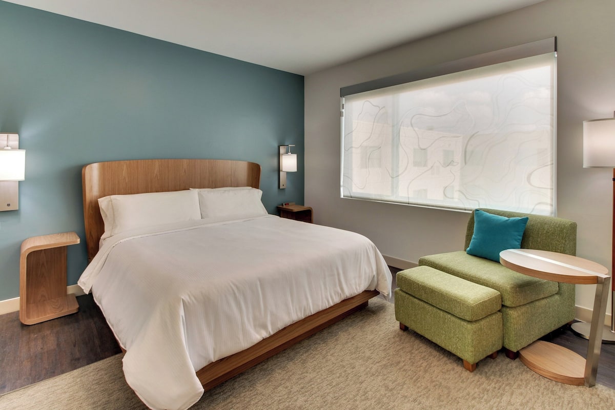 Quarto de hotel com uma cama, uma poltrona verde, uma mesa pequena e uma janela com vista para edifícios.