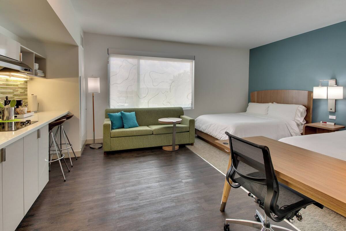 Quarto de hotel moderno com cama de madeira, sofá verde, mesa redonda pequena, escrivaninha com cadeira e área de kitchenette.