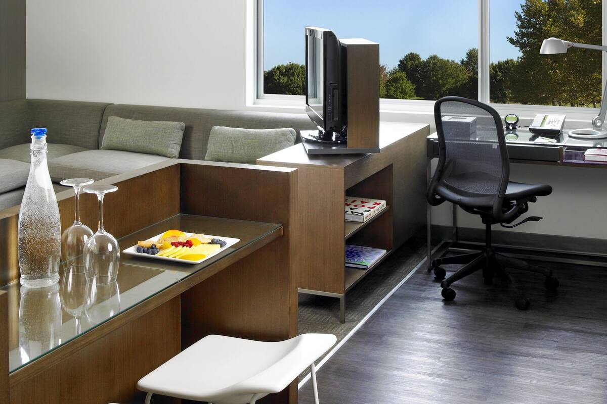 Uma sala de estar moderna com um sofá, uma mesa de vidro com uma garrafa e copos, uma televisão, uma escrivaninha com uma cadeira e uma grande janela com vista para árvores.