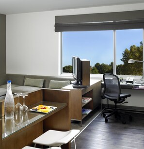 A modern living room with a sofa, a glass table with a bottle and glasses, a TV, a computer desk with a chair, and a large window with a view of trees.