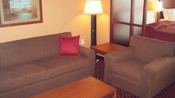 A hotel room with a sofa, armchair, and a small table. There is a lamp on a side table and a painting on the wall.
