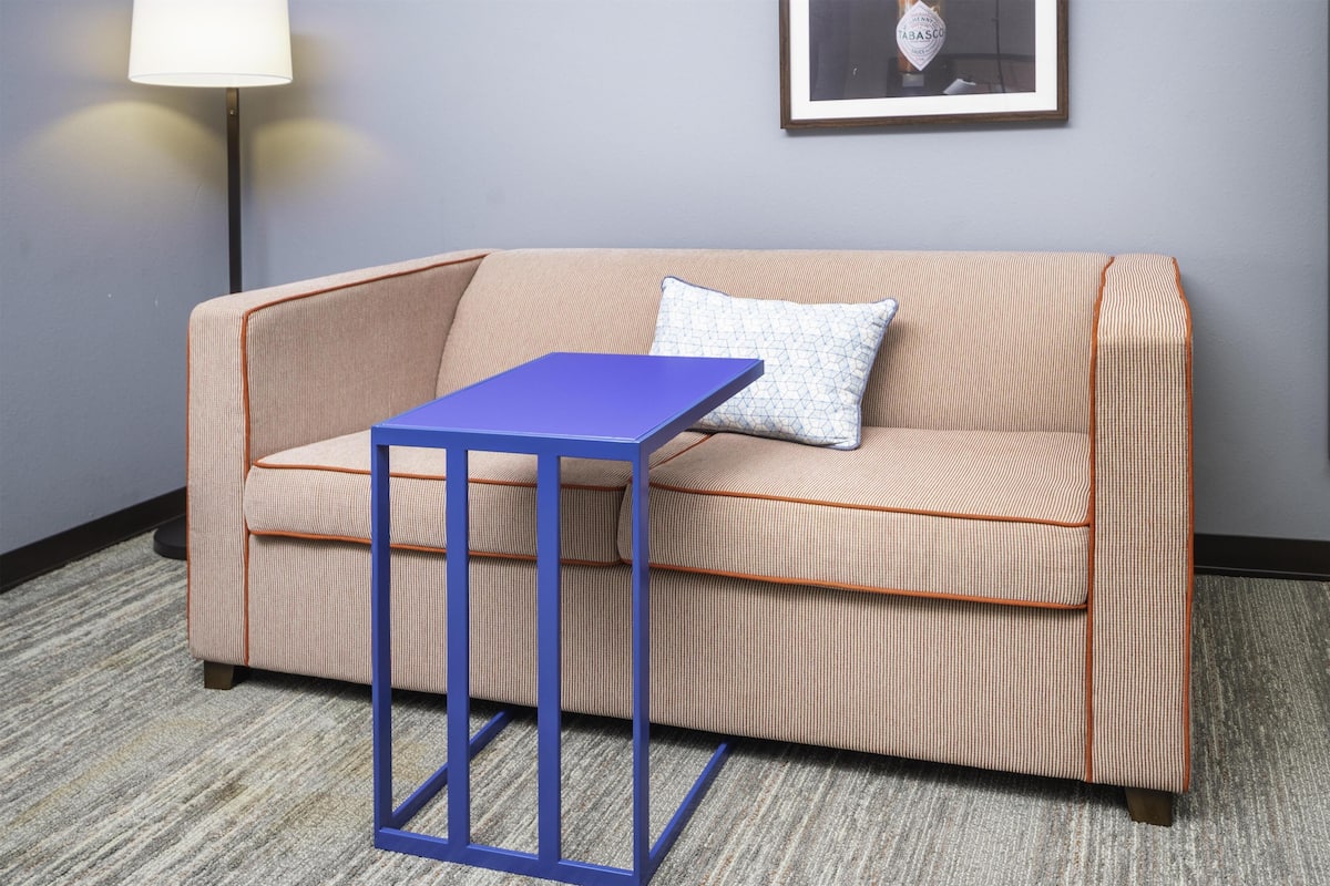 Una habitación con un sofá color beige, una mesita pequeña azul, un cojín estampado, un cuadro en la pared y una lámpara de pie.