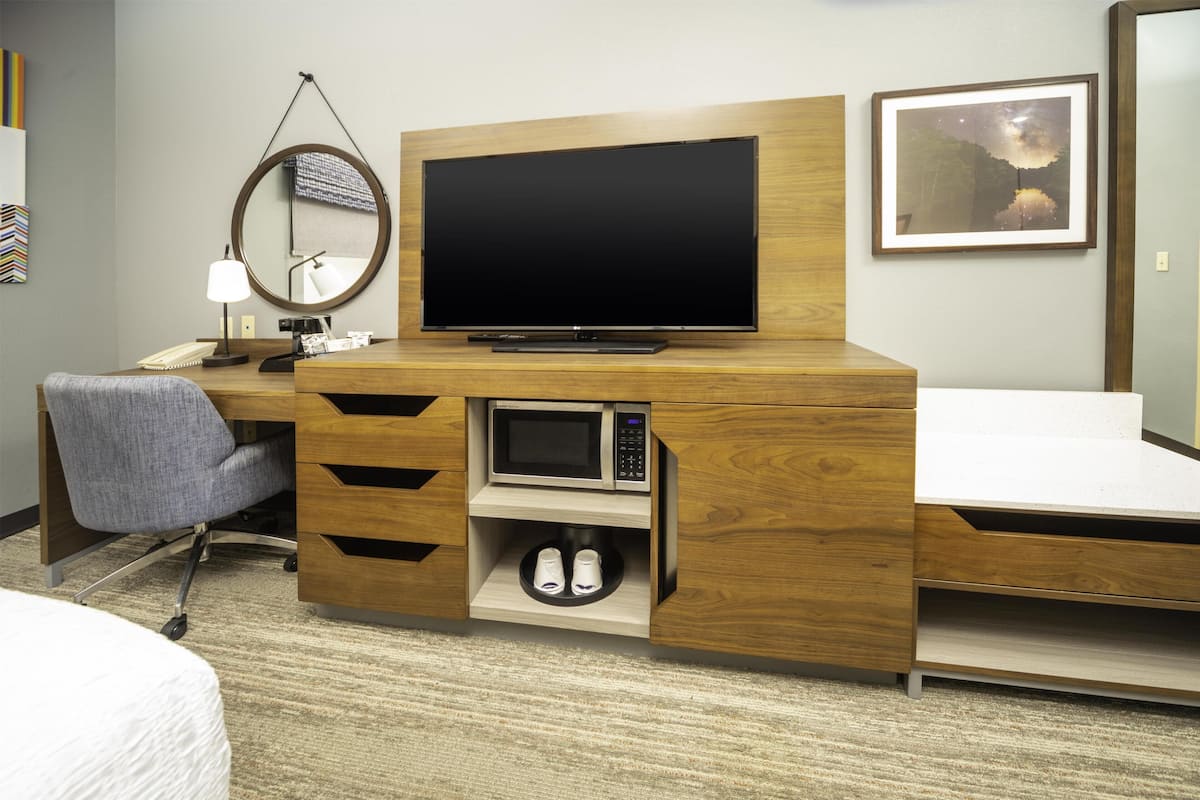 Una habitación de hotel con un escritorio de madera, televisión, espejo, silla, microondas y cama.