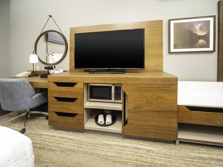 Quarto de hotel com escrivaninha de madeira, televisão, espelho, cadeira, micro-ondas e uma cama.