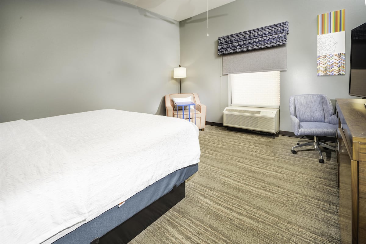 Habitación de hotel con cama, escritorio, silla, lámpara, ventana con persianas y televisor montado en la pared.