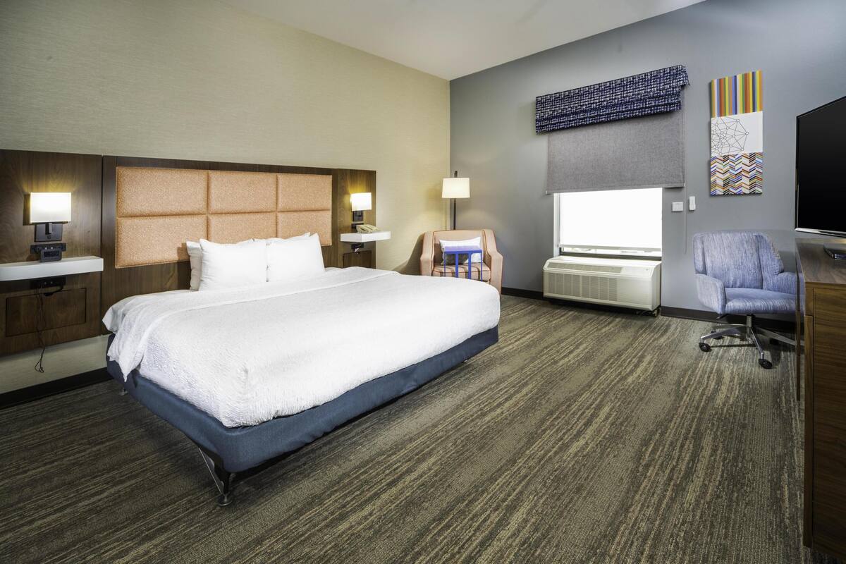 Habitación de hotel con una cama grande, dos mesitas de noche con lámparas, un escritorio con silla, un televisor de pantalla plana y una obra de arte en la pared.