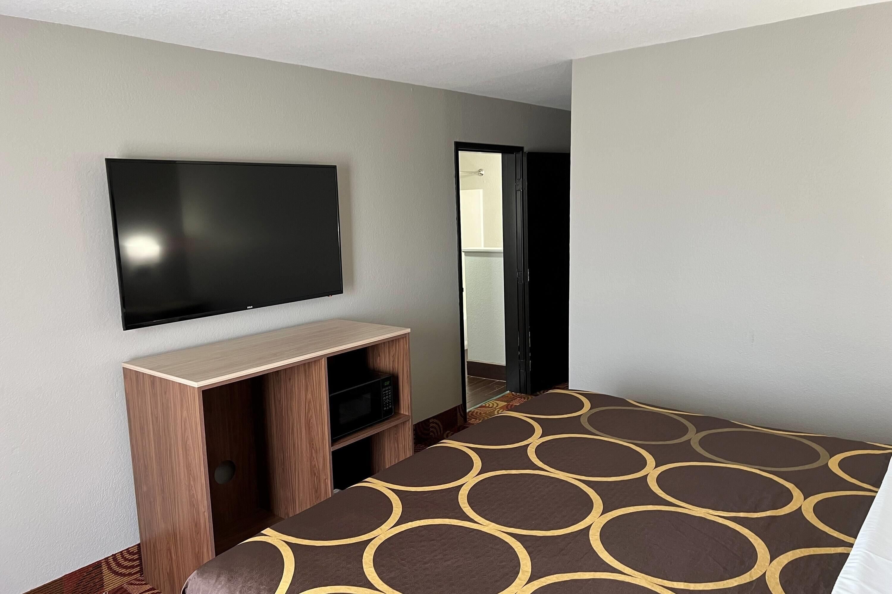 A hotel room with a flat-screen TV mounted on the wall, a wooden entertainment unit below, a bed with a patterned bedspread, and a door leading to a bathroom.