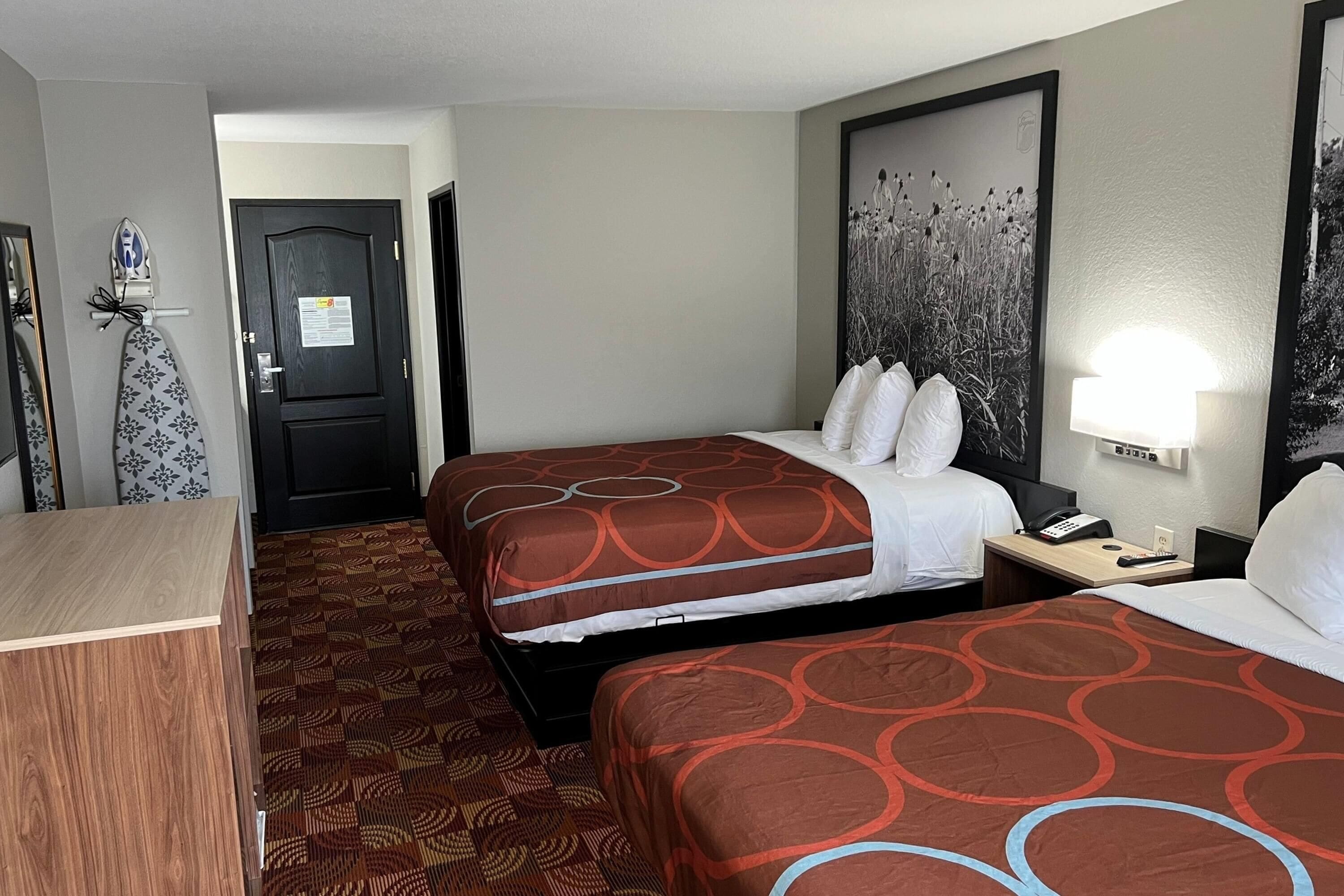 Quarto de hotel com duas camas, uma escrivaninha, uma cadeira, uma porta e uma luminária fixada na parede.
