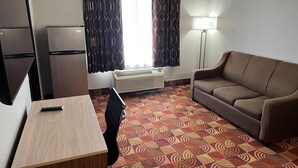 A compact hotel room with a kitchenette, a sofa, a small desk, and a patterned carpet.