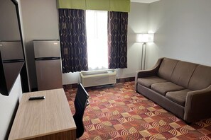 A compact hotel room with a kitchenette, a sofa, a small desk, and a patterned carpet.