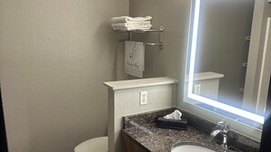 Baño con inodoro, lavabo y espejo. Hay un perchero con toallas dobladas y un letrero que dice 'Bolsa de Lavandería'.
