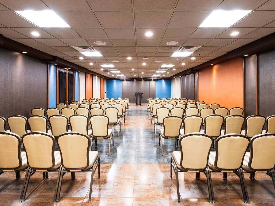A conference room with rows of chairs facing a podium, equipped with ceiling lights and a tiled floor.