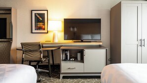 Una camera d'hotel con un letto, una scrivania con televisore, una sedia, un mobiletto con macchina del caffè e un quadro appeso al muro.
