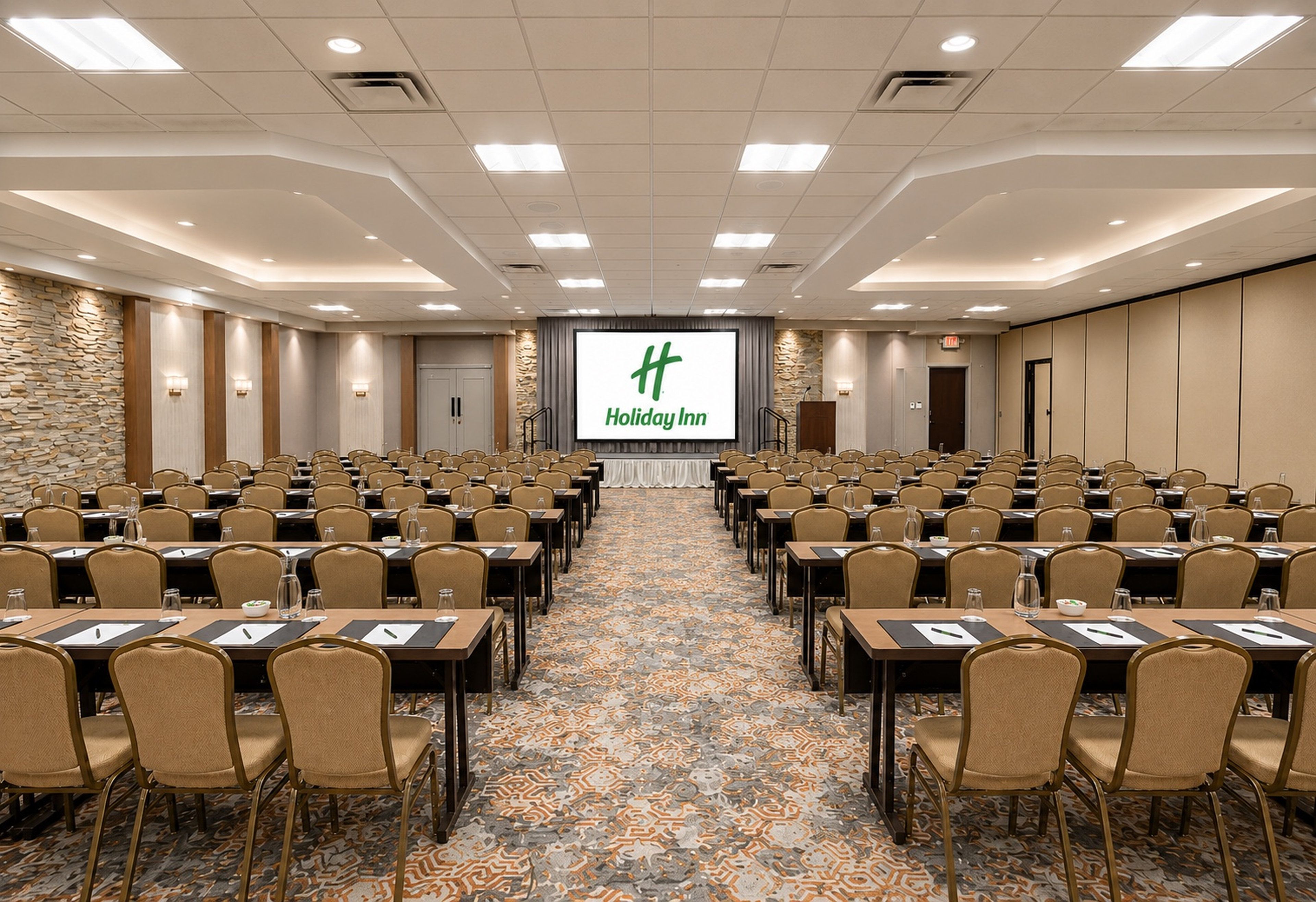 A conference room with rows of tables and chairs, a large screen at the front, and a patterned carpet.