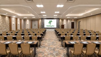 Un salón de conferencias con filas de mesas y sillas, una pantalla grande al frente y una alfombra estampada.