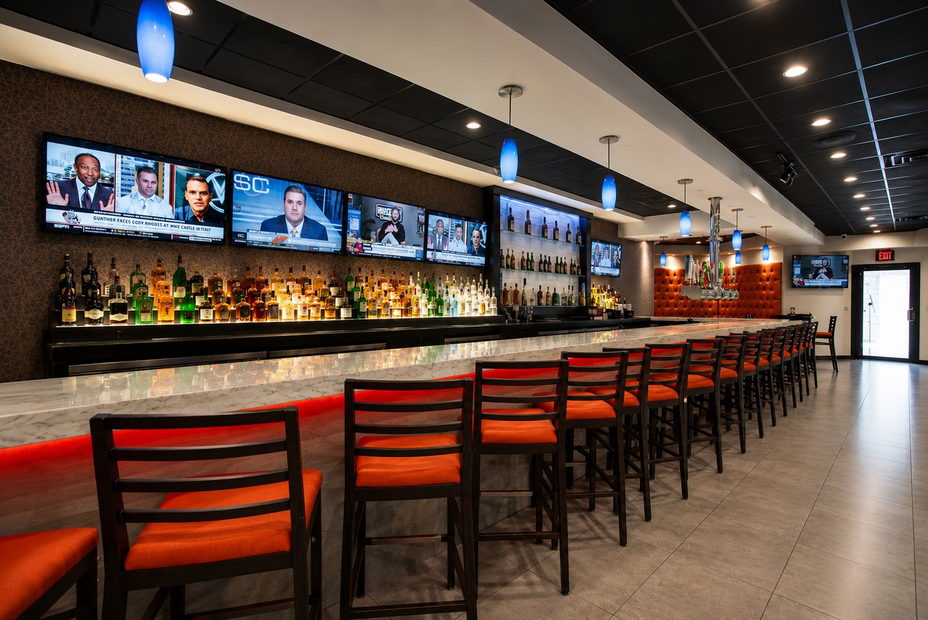 A bar area with multiple TV screens displaying news channels, a variety of liquor bottles, and bar stools with orange cushions.