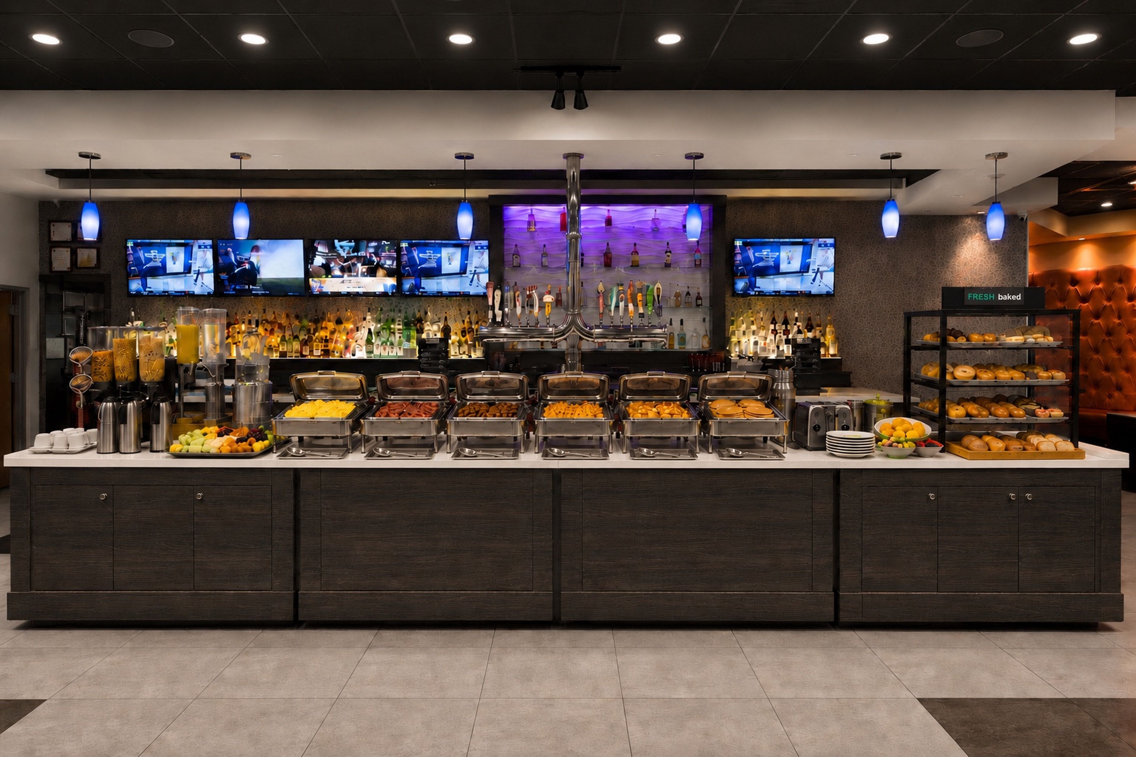 Zona de bufé con varias estaciones de comida, un bar con diversas bebidas y una pantalla de televisión.