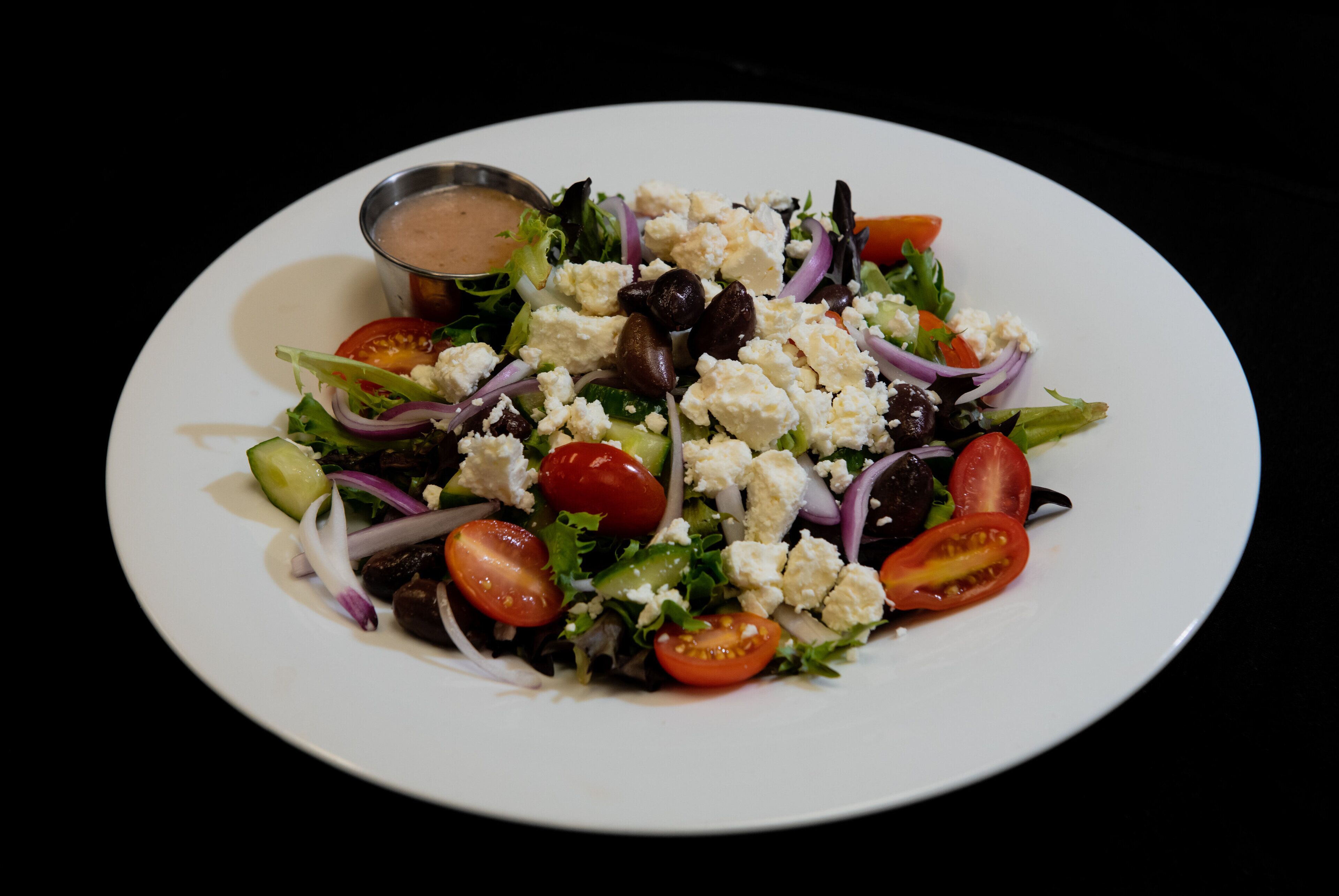 A plate of salad with various vegetables and feta cheese, accompanied by a small bowl of dressing.
