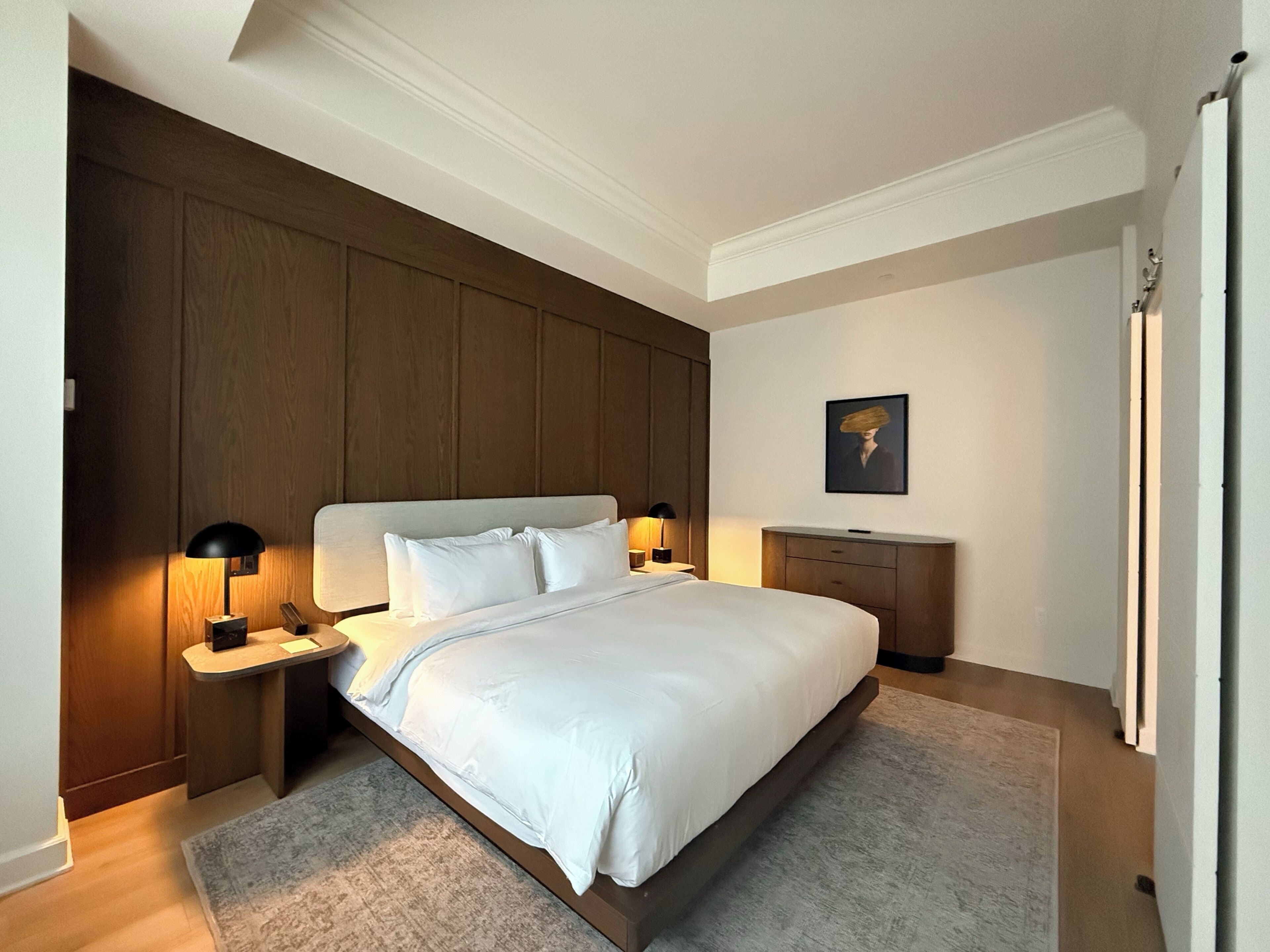 Une chambre d’hôtel avec un grand lit, deux tables de chevet avec des lampes, une tête de lit en bois, une petite table de chevet avec une lampe, un tableau encadré au mur et une porte avec un porte-serviettes.