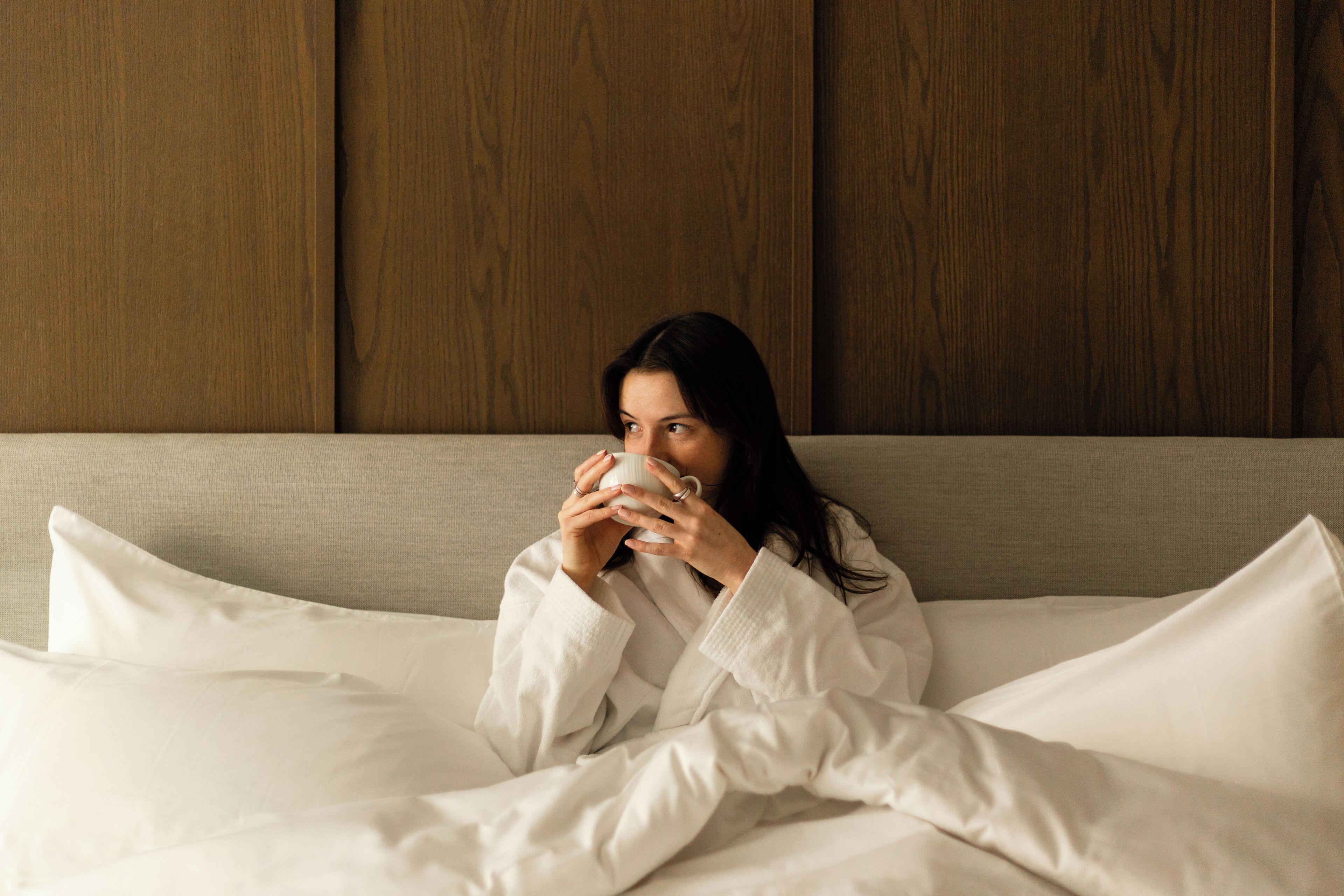 A person sitting on a bed with white bedding, a wooden headboard, and a plain wall in the background.