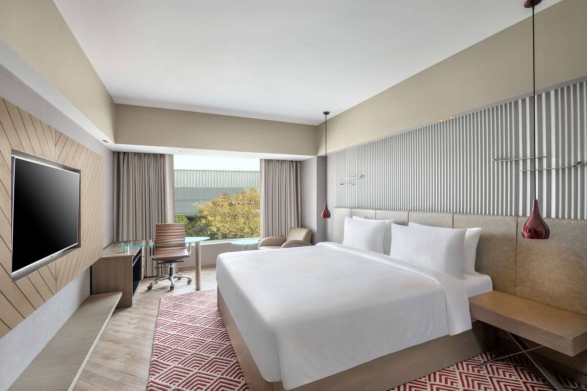 A modern hotel room with a large bed, a flat-screen TV, a desk, and a view of the outdoors.