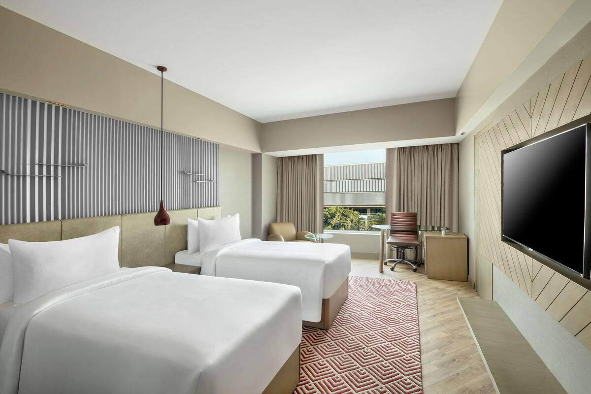 A hotel room with two beds, a TV, a desk, and a balcony with curtains.