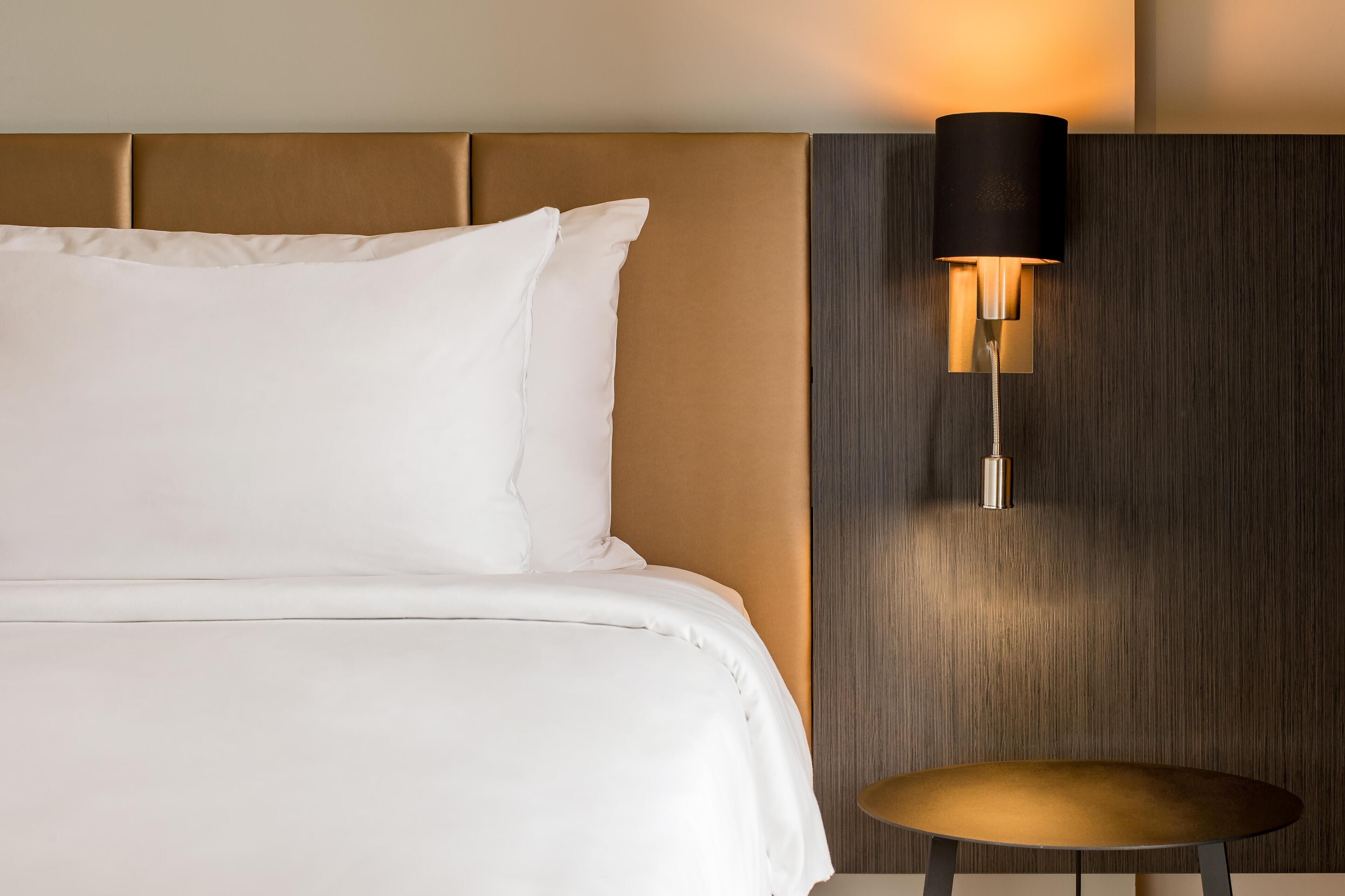 Cama bem arrumada com lençóis brancos, mesa redonda com tampo dourado e luminária fixada na parede.