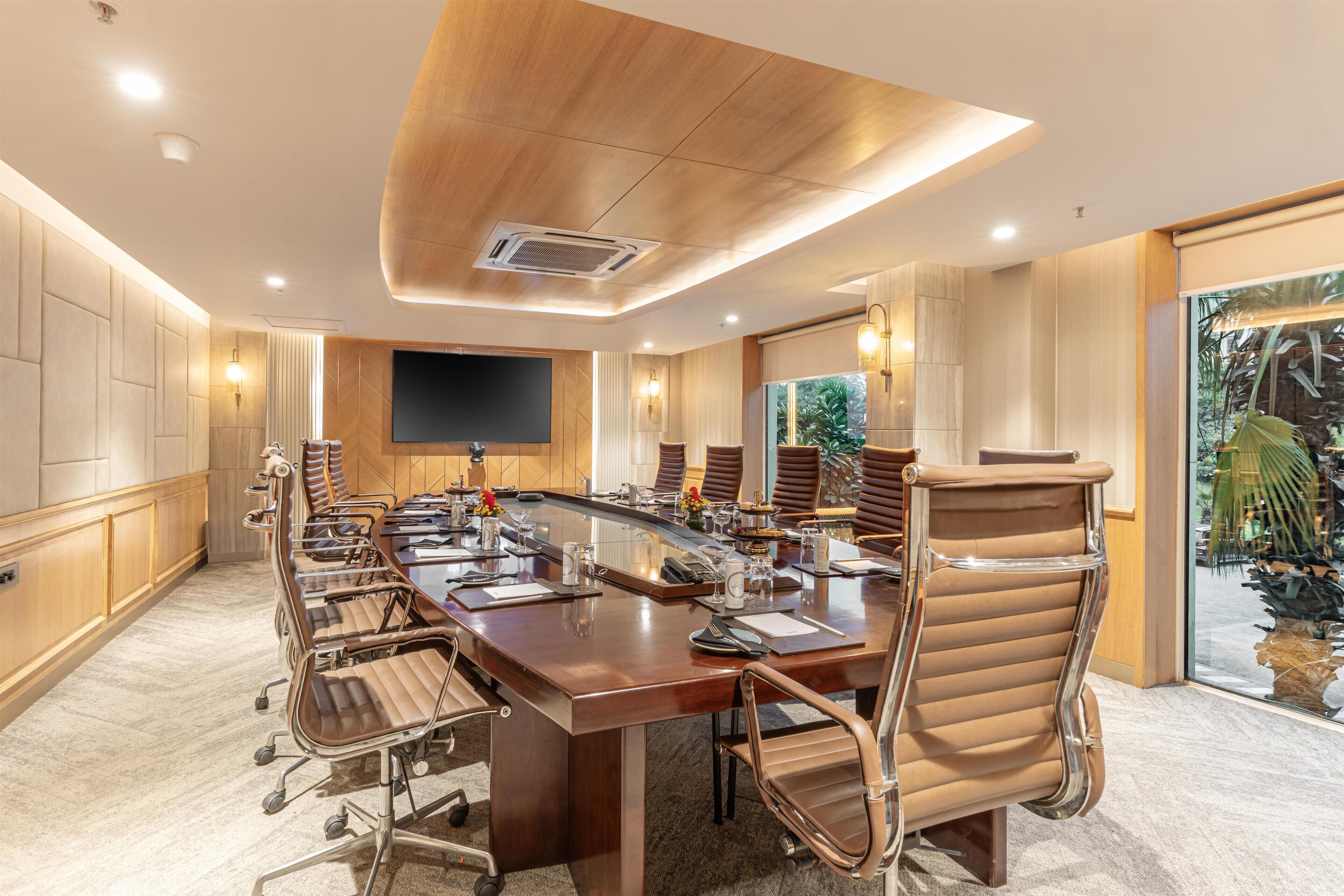 A conference room with a long table, chairs, a flat-screen TV, and a large window with a view of greenery.