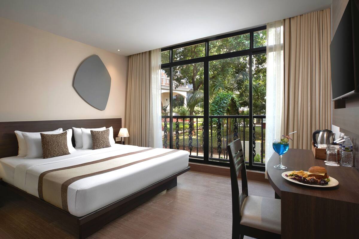 A hotel room with a large bed, a desk with a plate of food and a drink, and a view of greenery outside.
