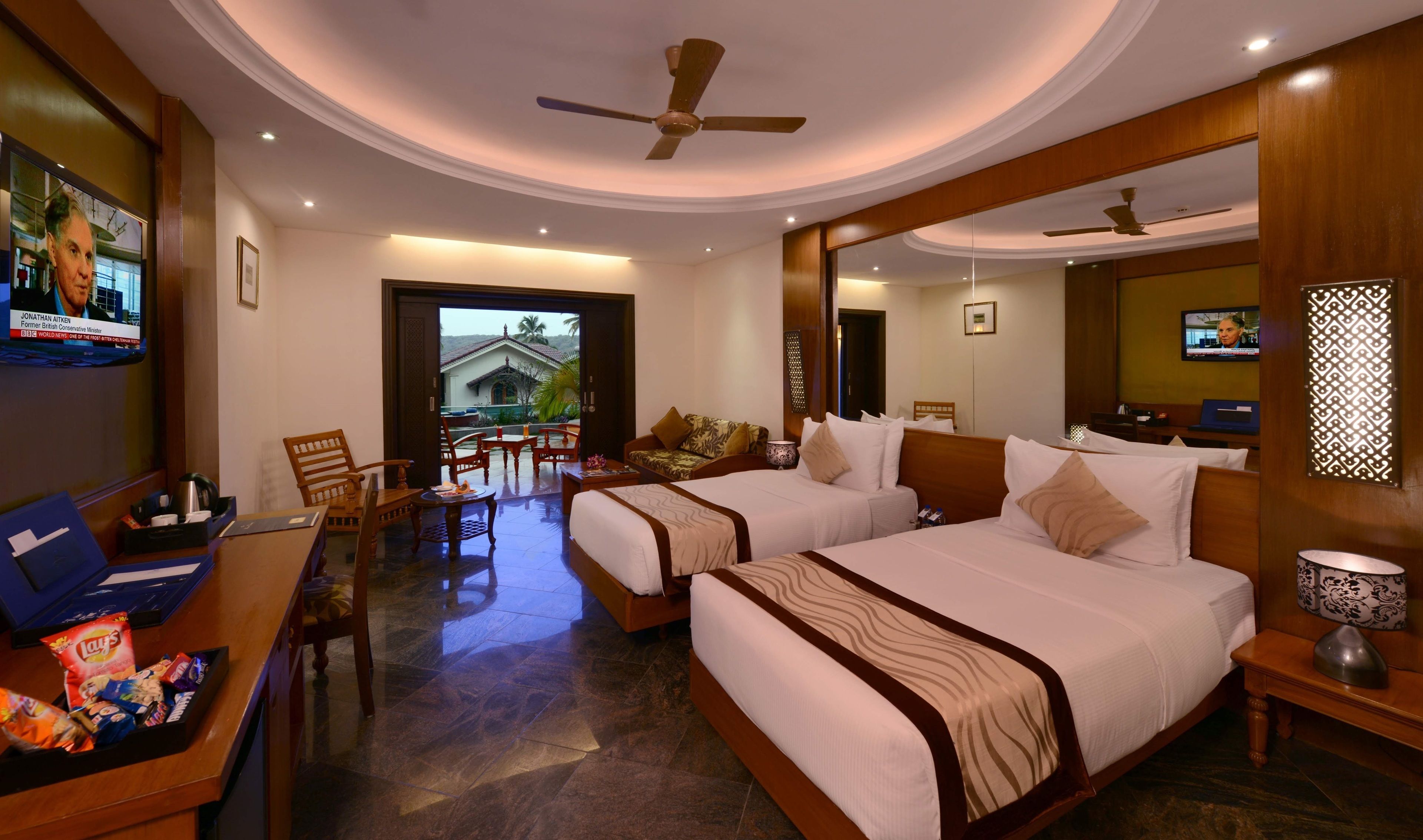 A hotel room with two beds, a desk with snacks, a TV, and a balcony with a view of a house.