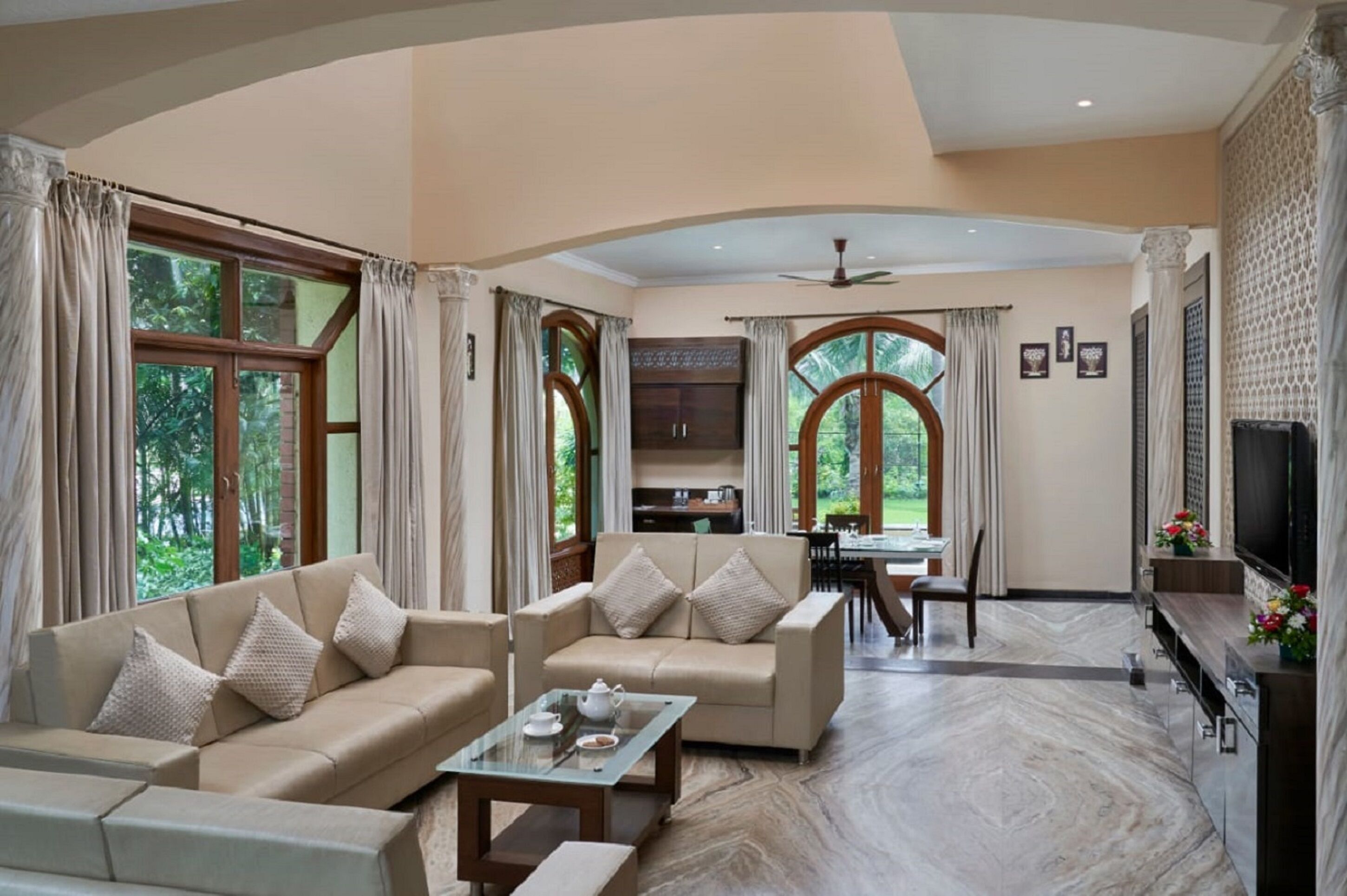 A spacious living area with a sofa set, a coffee table, and a TV. There are large windows with curtains, a ceiling fan, and a dining area in the background.