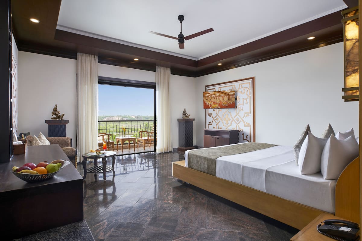 A modern hotel room with a large bed, a balcony with a view, a dining area, and a fruit bowl on the side table.